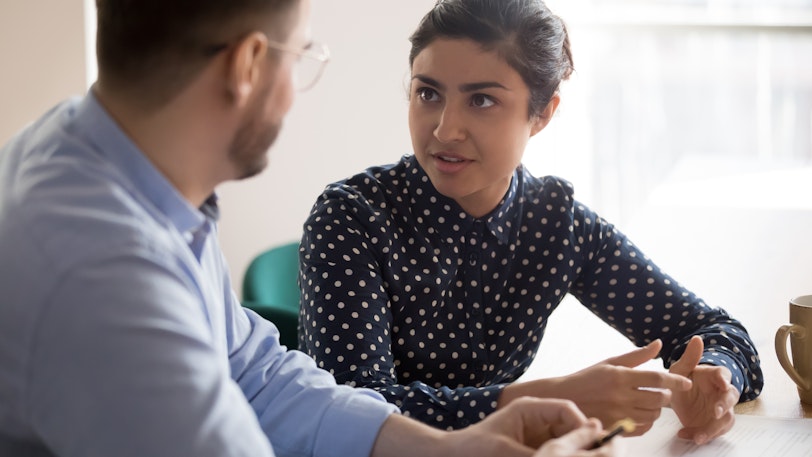 Young indian female mentor coach worker talking to male coworker teaching intern having business conversation with workmate, serious hindu manager helping colleague discussing new project in office