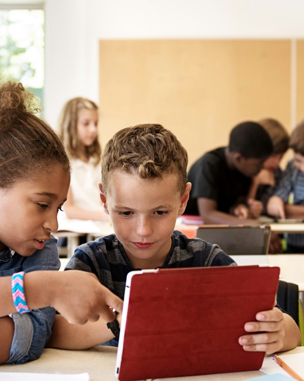 Two young students sitting together in a classroom and looking at a tablet screen with a red protective cover on it.